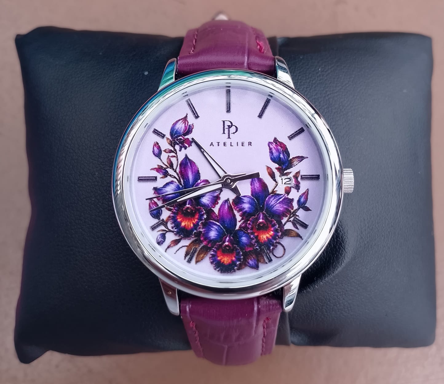 PP Atelier Cattleya em seu estojo, com caixa redonda prateada, mostrador lilás com orquídeas, janela de data e pulseira roxa.